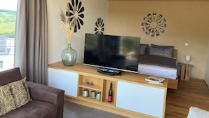 A modern living room with a TV, wooden furniture, decorative wall art, and a view of greenery outside. - MoselHoch vacation cottage in the vineyards of Klüsserath (Klüsserath)