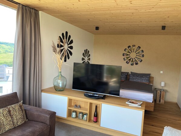 A modern living room with a TV, wooden furniture, decorative wall art, and a view of greenery outside.