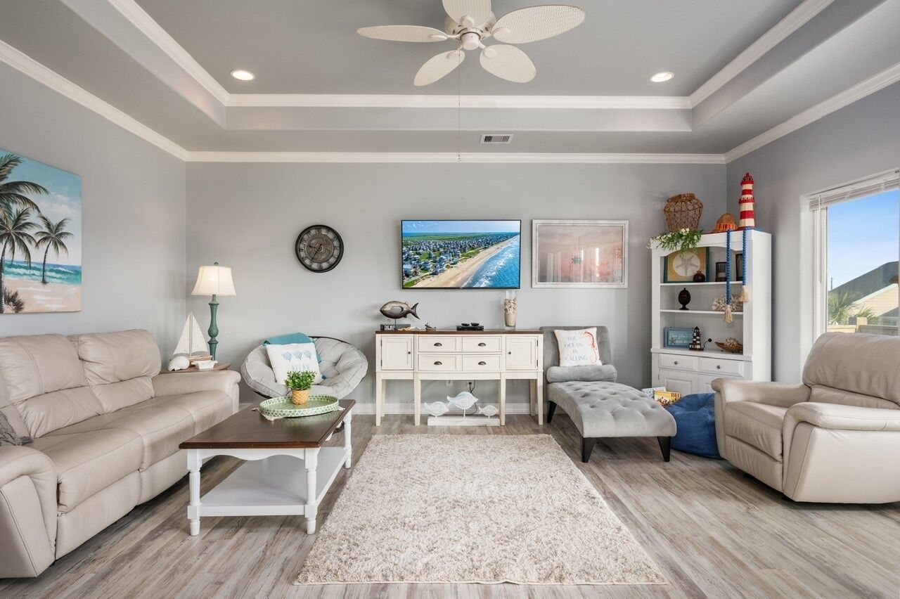 Una sala de estar con un sofá color beige, una mesa de centro blanca, un sillón gris, una mesita de noche blanca con una lámpara, un mueble consola blanco con adornos, un televisor de pantalla plana grande y una ventana con vista a la playa.