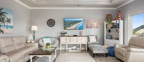 Una sala de estar con un sofá color beige, una mesa de centro blanca, un sillón gris, una mesita de noche blanca con una lámpara, un mueble consola blanco con adornos, un televisor de pantalla plana grande y una ventana con vista a la playa.