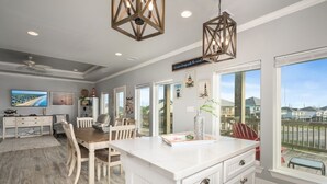 A spacious kitchen with a large island, a dining area with wooden chairs, and a view of the neighborhood through large windows. - Big Chill 2 Bedroom Home (Port Bolivar)