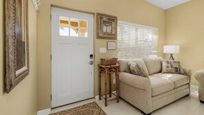 A living room with a beige sofa, a white door, a window with blinds, a wall-mounted picture frame, and a small table with a basket.