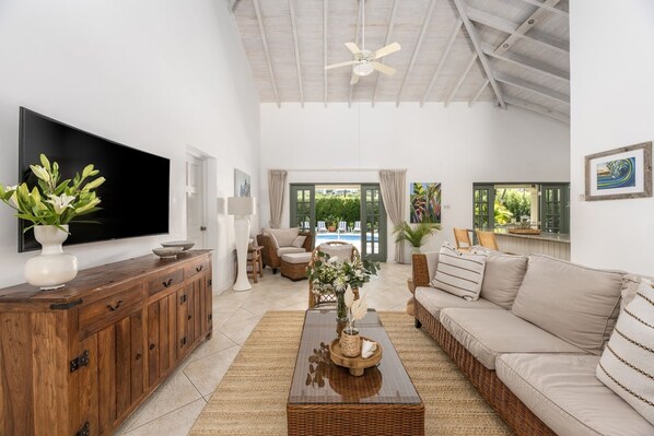 A spacious living room with a large flat-screen TV, a wooden cabinet, a wicker sofa, and a view of a pool through the open door. - Gibbs Glade Villa 3 Home (Gibbes)
