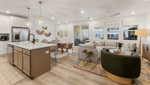 Et moderne køkken med en central ø, spiseområde med fletstole, opholdsstue med sofa og sofabord, samt store vinduer med udsigt.
