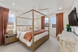A four-poster bed with a wooden frame, a bedside table with drawers, a lamp, and a view of the city through large windows. - Utah Utopia 3 Bedroom Condo (St. George)