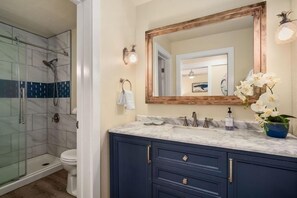 Ein Badezimmer mit Duschkabine aus Glas, einer weißen Toilette, einem мраморный Waschbecken mit einem spiegel mit Holzrahmen und einem blauen Schrank.