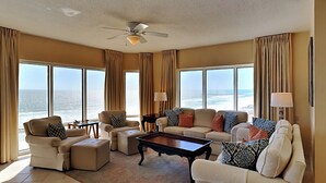 A spacious living room with a large window overlooking the ocean, furnished with a sofa, chairs, and a coffee table. - Emerald Isle | Corner Condo w. Pool/Hot Tub, Private Beach + More (Pensacola Beach)
