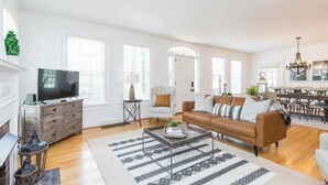 Amplia sala de estar con un sofá de cuero marrón, una mesa de centro y un televisor sobre un mueble de madera. Cuenta con un comedor con una araña y una chimenea.