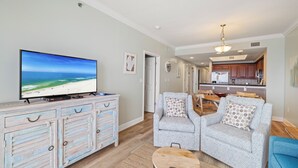 Un salon avec un téléviseur, deux fauteuils gris, une table basse et, au fond, une cuisine.