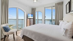 A spacious bedroom with a large bed, a TV, and a view of the sea through large windows.