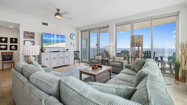 A spacious living room with a large sofa, a coffee table, and a television. There is a view of the ocean through large windows.
