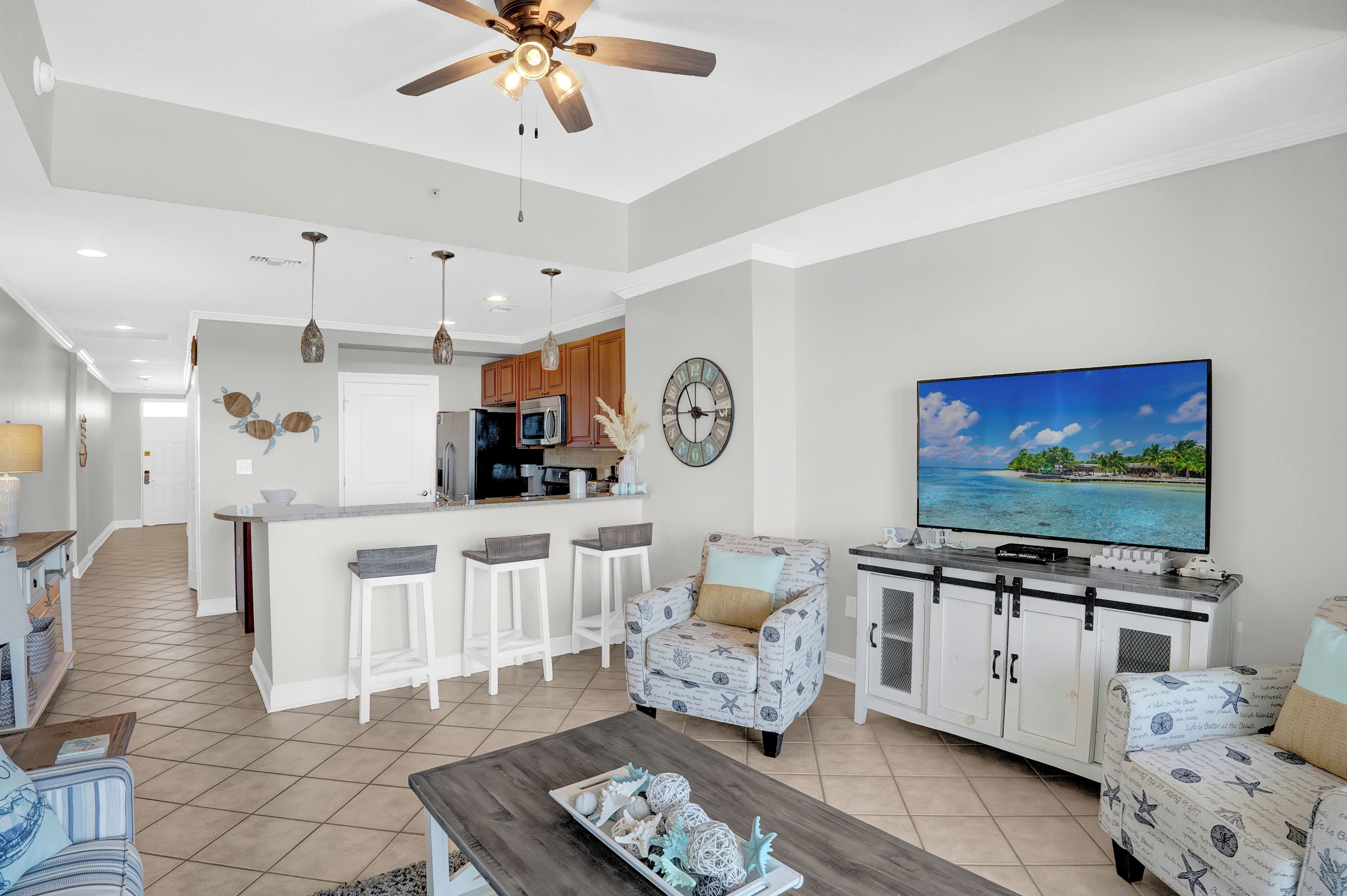 A modern living room with a TV, a ceiling fan, and a kitchen area in the background.