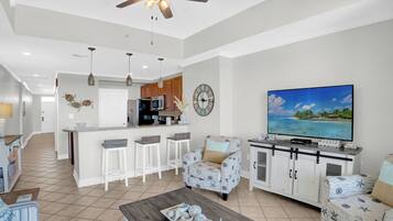A modern living room with a TV, a ceiling fan, and a kitchen area in the background.