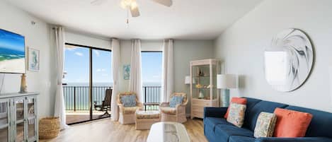 A spacious living room with a glass coffee table, a blue sofa, wicker chairs, and a view of the ocean through large windows.