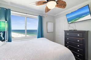 A bedroom with a large bed, a dresser, a television, and a view of the beach.
