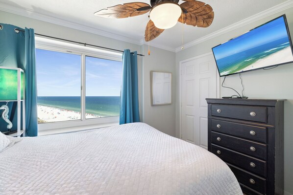 A bedroom with a large bed, a dresser, a television, and a view of the beach.