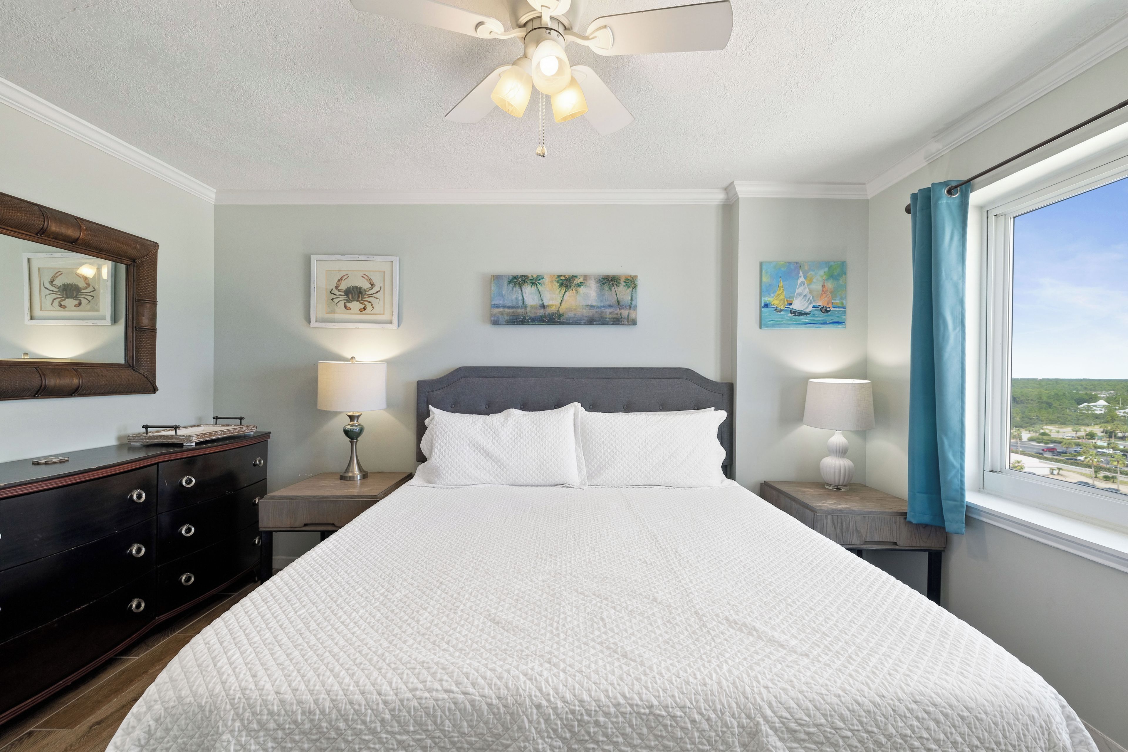 A bedroom with a large bed, a ceiling fan, a nightstand with a lamp, a dresser, and artwork on the walls.