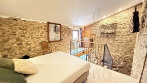 A bedroom with a stone wall, a bed, a chair, a small table, and a balcony with a railing. - Agi Porthos 800 Apartamento for rent in Roses Girona. (Roses)