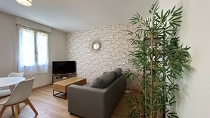 Un salon avec un mur en pierre, un téléviseur à écran plat, un canapé gris, une table à manger avec des chaises et une grande plante en pot.