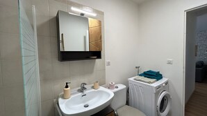 Une petite salle de bain avec un lavabo blanc, un miroir et des toilettes. On peut apercevoir un lave-linge dans un coin.