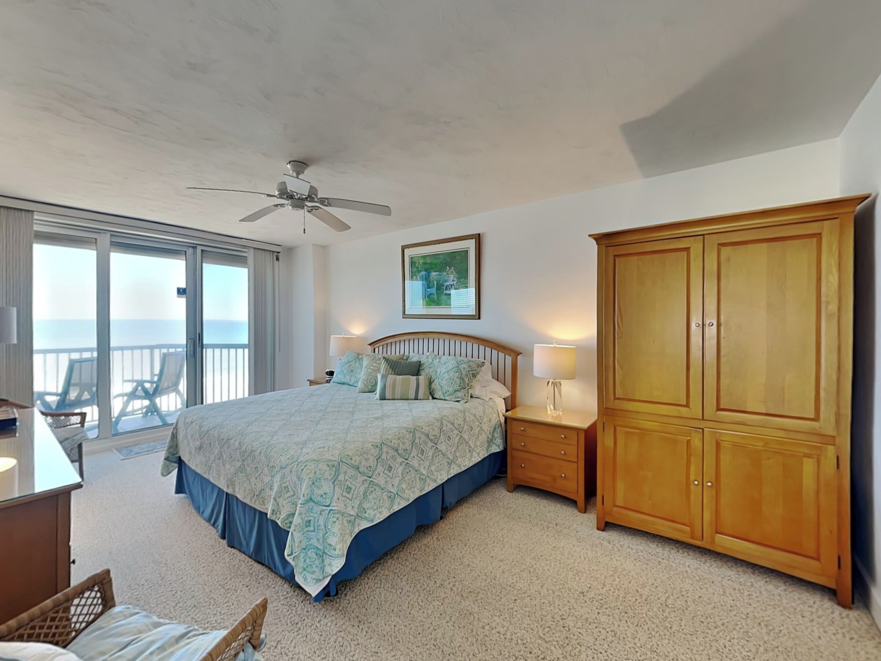 Une chambre à coucher comprenant un lit, une table de chevet, une armoire en bois, un ventilateur de plafond et un balcon avec des chaises.