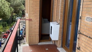 Een balkon met uitzicht op geparkeerde auto's en bomen, een wit apparaat op de vloer, en een deur die naar een binnenruimte leidt.