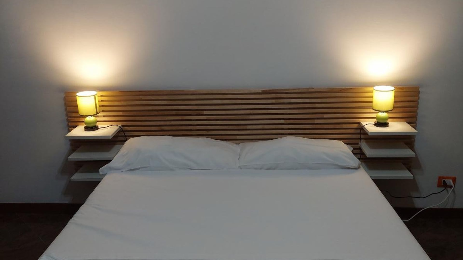 Un lit recouvert de draps blancs, flanqué de deux tables de chevet surmontées de lampes, adossé à un mur orné de lattes de bois horizontales.