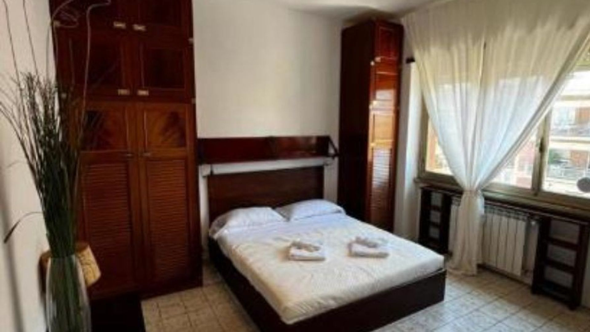 Un dormitorio con cama, armario, radiador y ventana con cortinas.