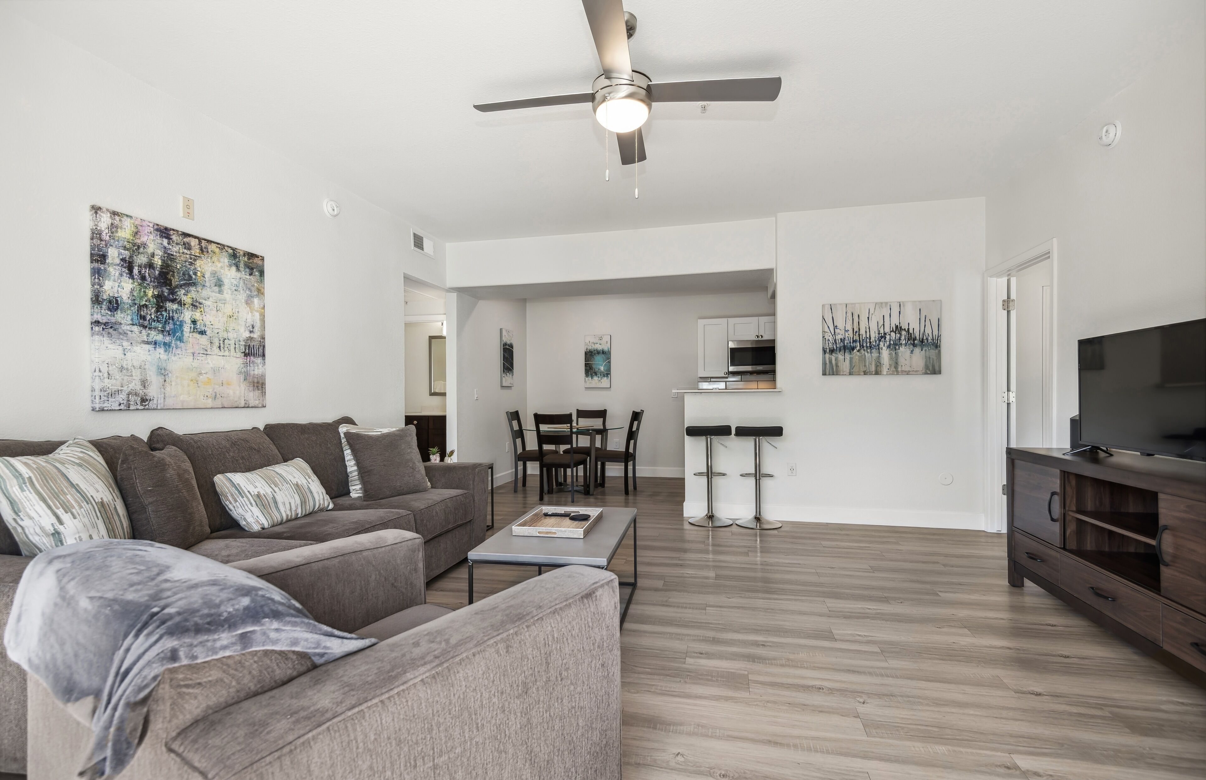 A modern living room with a grey sofa, a coffee table, and a TV stand. There is a dining area with a table and chairs in the background.