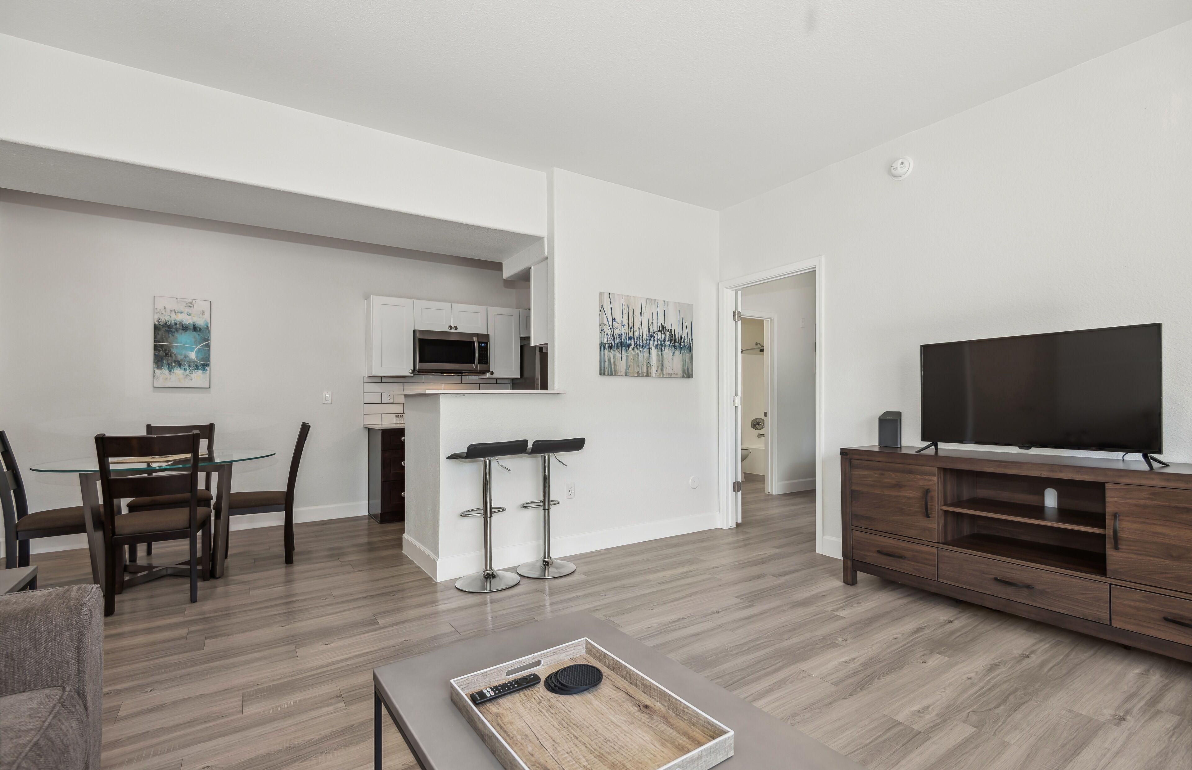 A modern living room with a TV, wooden flooring, a dining area with chairs, and a kitchen in the background.