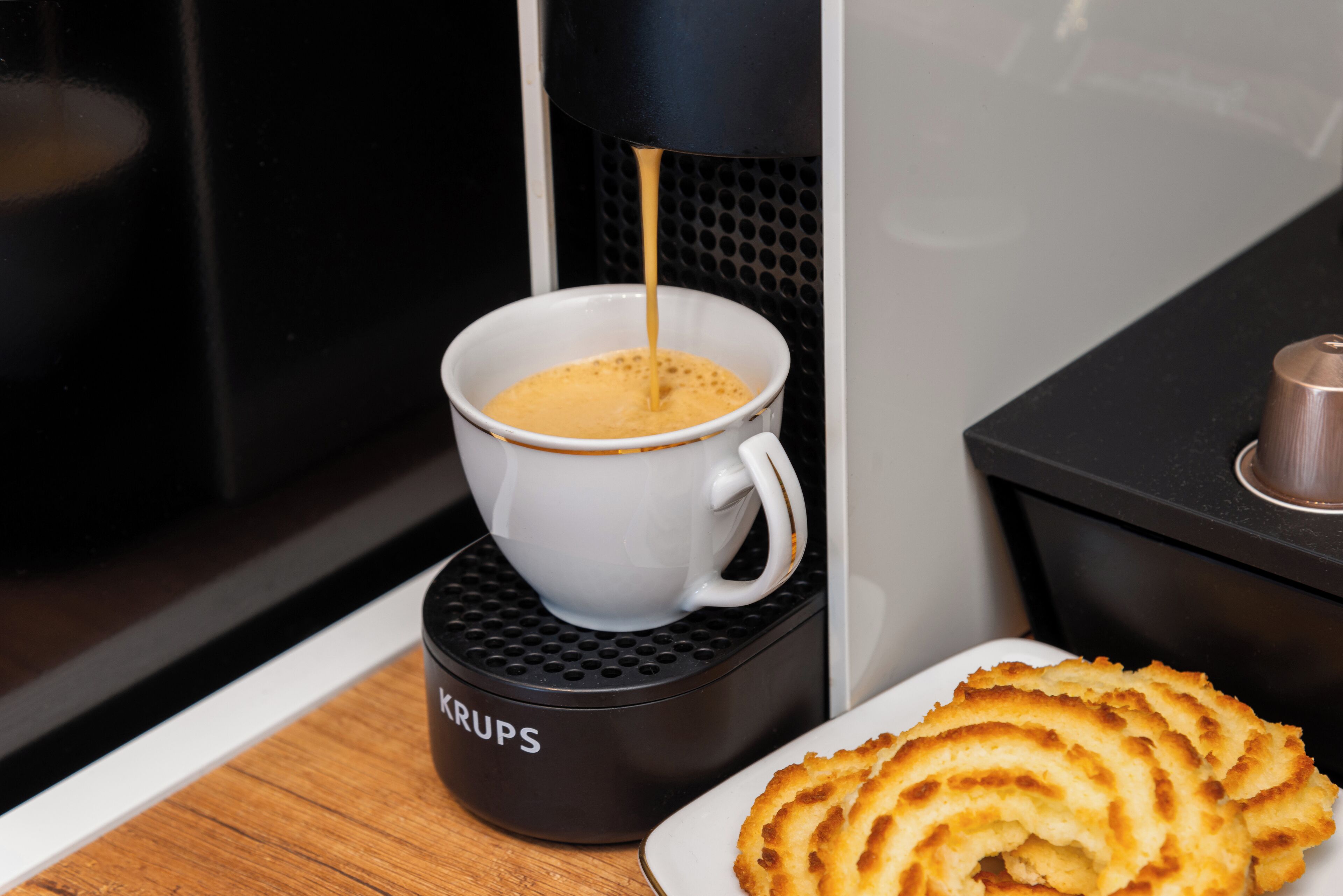 Une machine à café qui verse du café dans une tasse, une assiette de biscuits et une cafetière noire sur une table en bois.