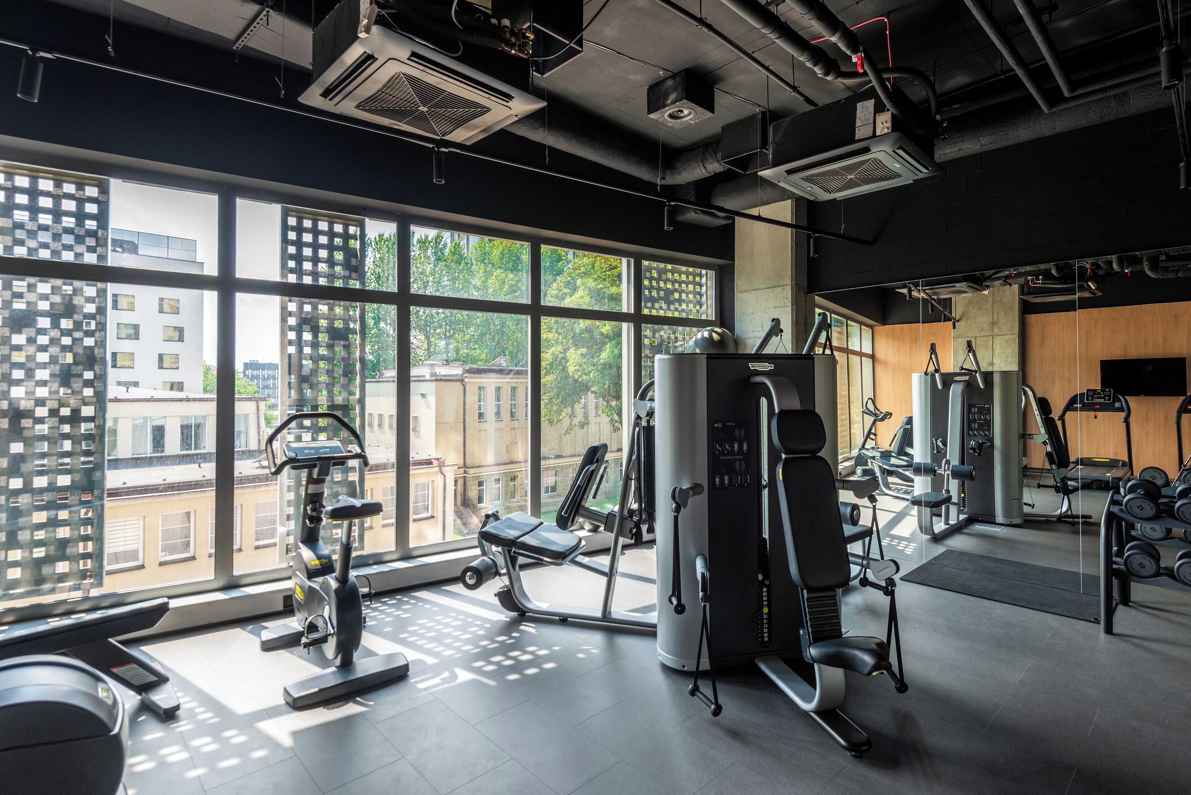 A gym with various exercise equipment, large windows, and a view of buildings outside.