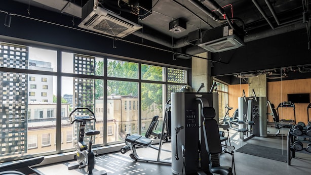 Et fitnesscenter med forskellig træningsudstyr, store vinduer og udsigt til bygninger.