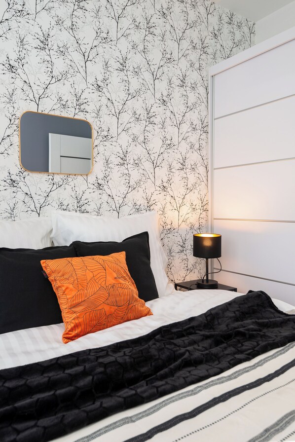 Un lit bien fait, avec une literie noire et blanche, un coussin à motifs orange et une couverture noire texturée. Une lampe noire est posée sur une table de chevet, à côté du lit.