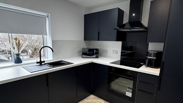A modern kitchen with black cabinets, a herringbone wood floor, a built-in oven, and a window with a view of trees.