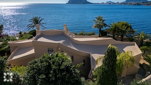 A Mediterranean-style villa with a curved roof, surrounded by lush greenery and overlooking a calm sea with a distant mountain and cityscape.