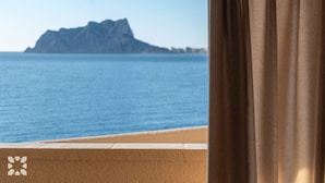 A view of a beach with a mountain in the distance, a balcony with a curtain, and a sandy area.