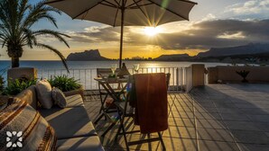 A terrace with a table, chairs, and an umbrella, overlooking a body of water and mountains during sunset.