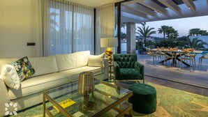 Ein modernes Wohnzimmer mit einer Couch, einem Glastisch, einem grünen Sessel und einem Essbereich mit Gartenmöbeln.