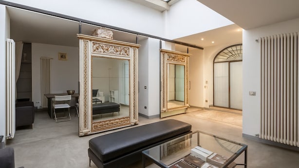 A spacious living area with a high ceiling, a skylight, a large ornate mirror, a black bench, and a glass coffee table. - dHomes -Loft suite in Duomo (Milan)