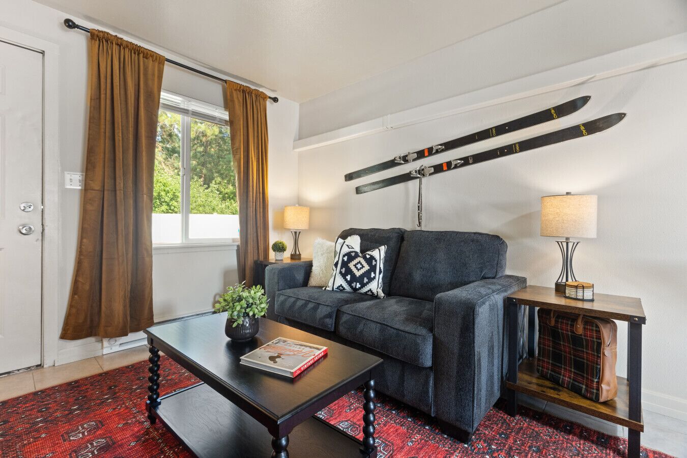 Une pièce comprenant un canapé, une table basse avec un livre, une lampe, un porte-manteau avec des paires de skis, et une fenêtre avec des rideaux.
