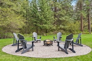 A circular seating area with four Adirondack chairs around a central fire pit, set on a gravel circle in a grassy field with trees in the background.