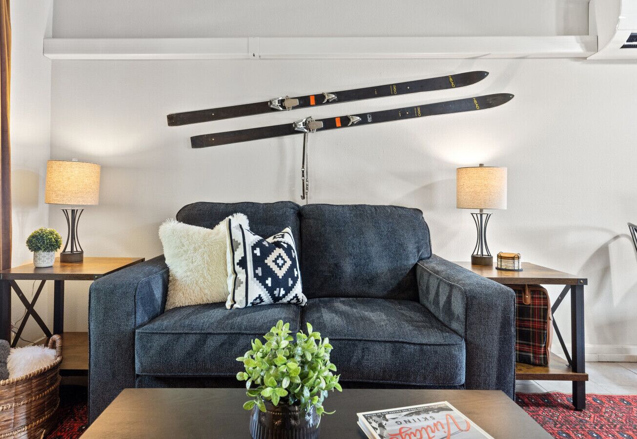 A modern living room with a sofa, two side tables with lamps, a coffee table with a plant, and a ski rack above the sofa.