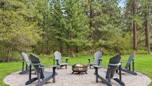 Ein kreisförmiger Sitzbereich mit vier Adirondack-Stühlen um eine zentrale Feuerstelle, auf einem Kiesrondell in einem grünen Feld mit Bäumen im Hintergrund.