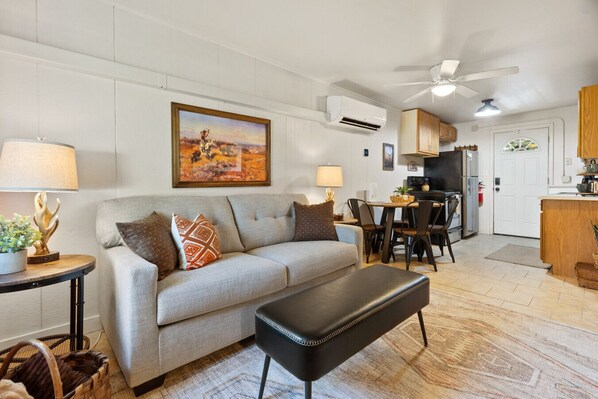 A living room with a sofa, a dining table with chairs, a black bench, a painting on the wall, and an air conditioning unit.
