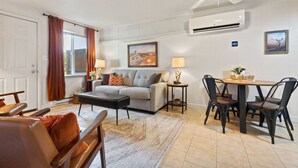 Ein Wohnzimmer mit einer Couch, einem Esstisch mit Stühlen, einem Deckenventilator, einer Klimaanlage und einem Fenster mit Vorhängen.