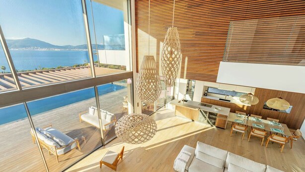 Interior moderno com uma janela ampla que oferece vista para a água, uma área de refeições com mobiliário de madeira e um espaço de estar com sofá e cadeiras.
