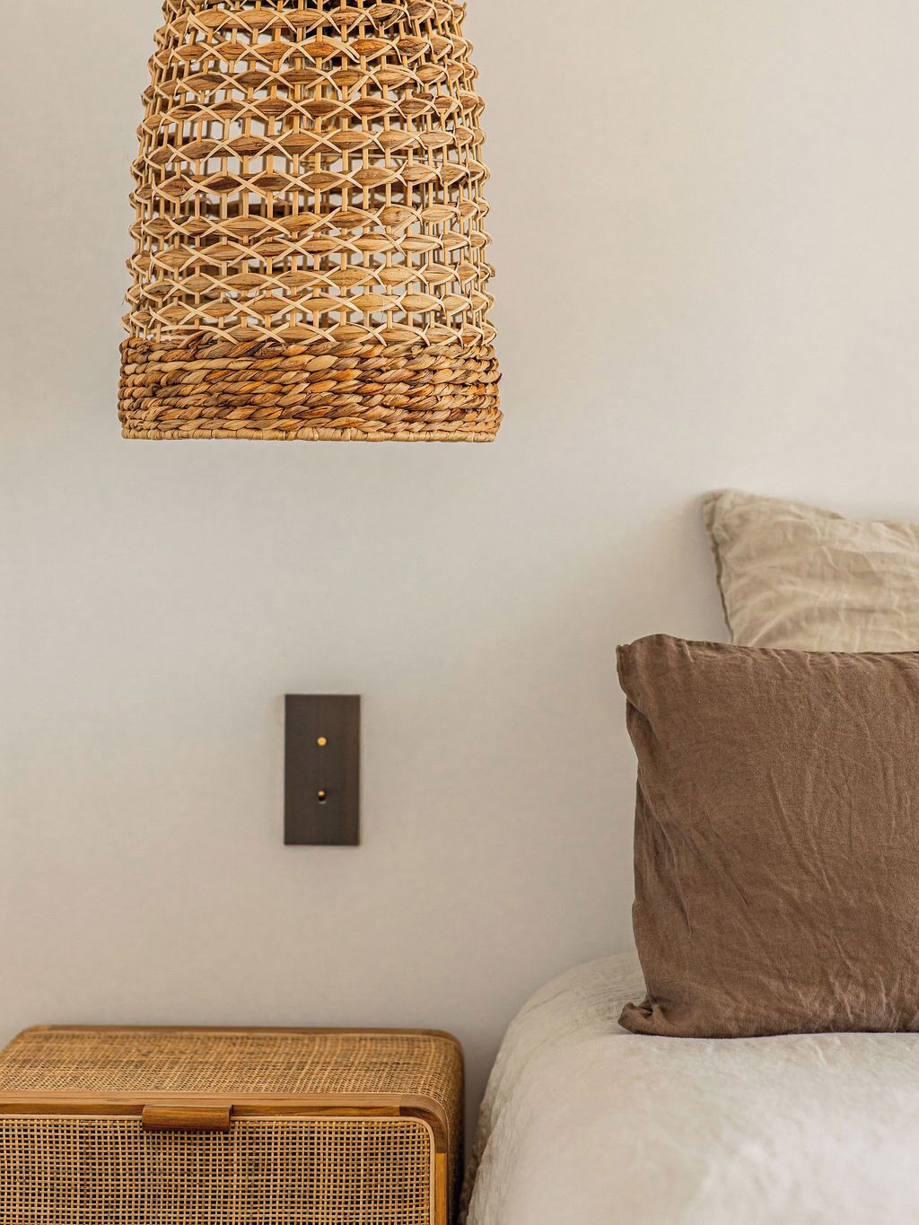 Une pièce avec une décoration murale en rotin tressé, une table de chevet, un lit avec un oreiller marron et un interrupteur fixé au mur.
