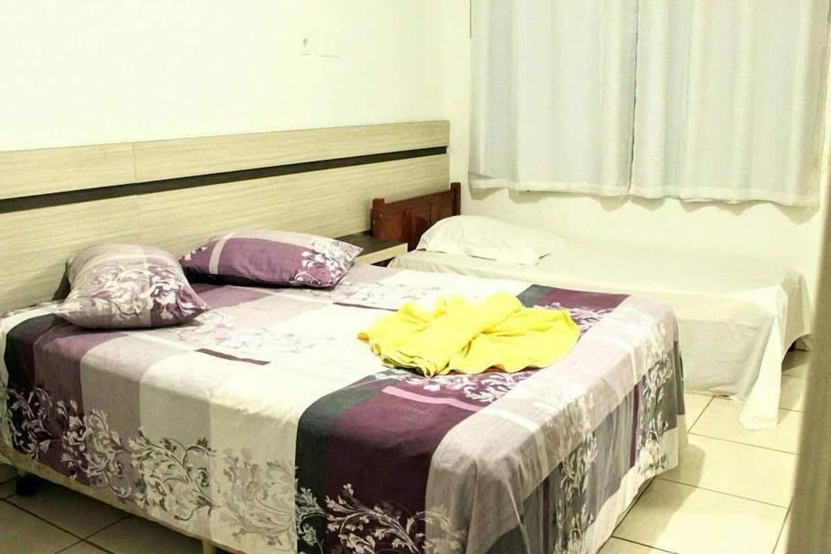 A single bed with a floral bedspread and two pillows, a bedside table with a yellow towel, and a window with white curtains.