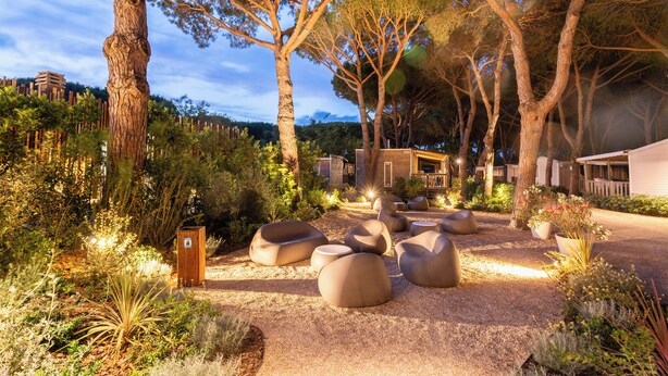 A landscaped garden with large trees, outdoor seating, and a building with a sloped roof. - Hotiday Lodge Orbetello (Orbetello)